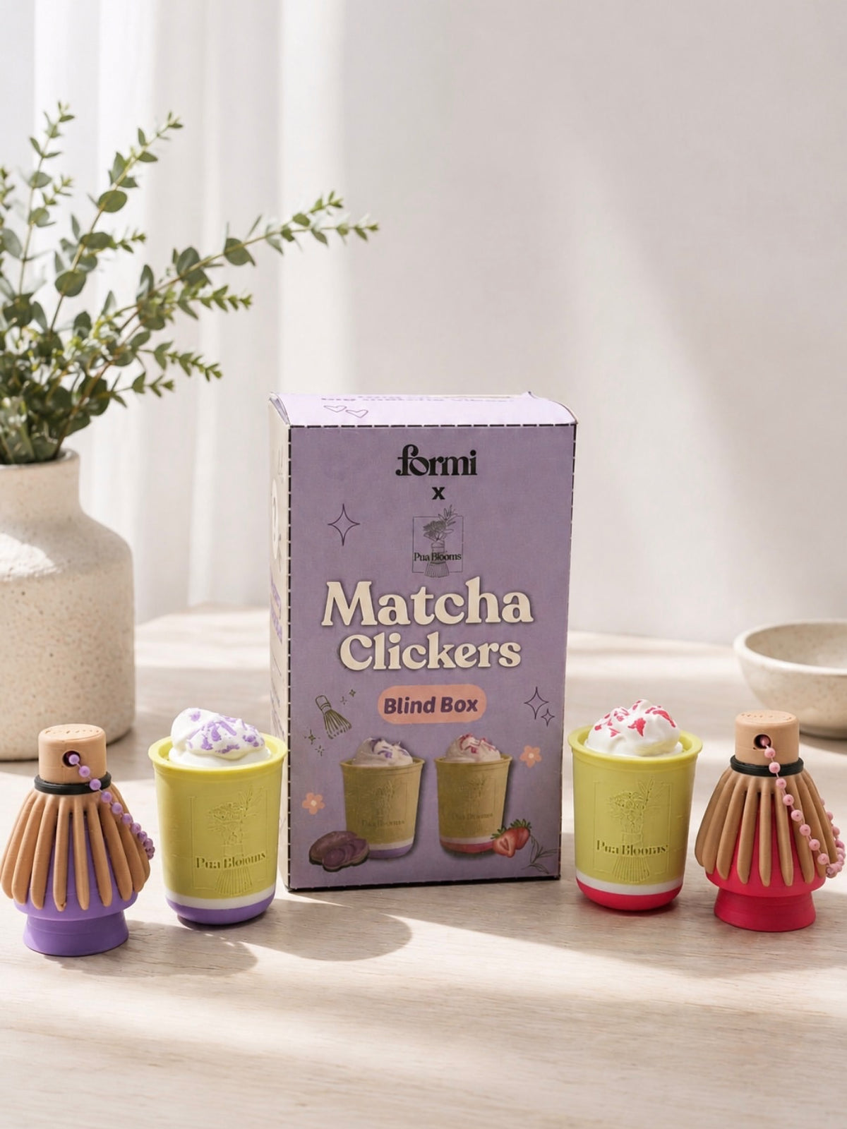 Formi x Pua Blooms matcha clickers and whisk keychains beside their lavender blind-box packaging
